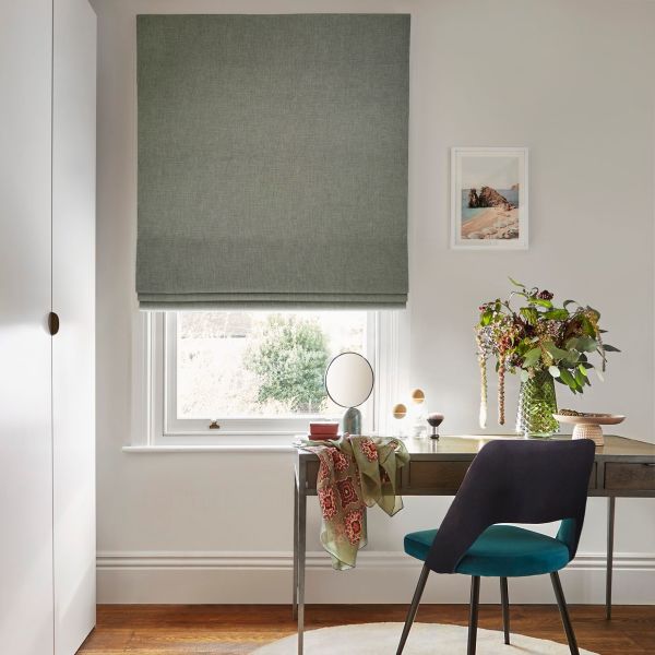 Green fabric Roman blind, lowered and lightly filtering daylight; small desk and teal chair beneath, vase of flowers and mirror on the desk, framed coastal print on a neutral wall.