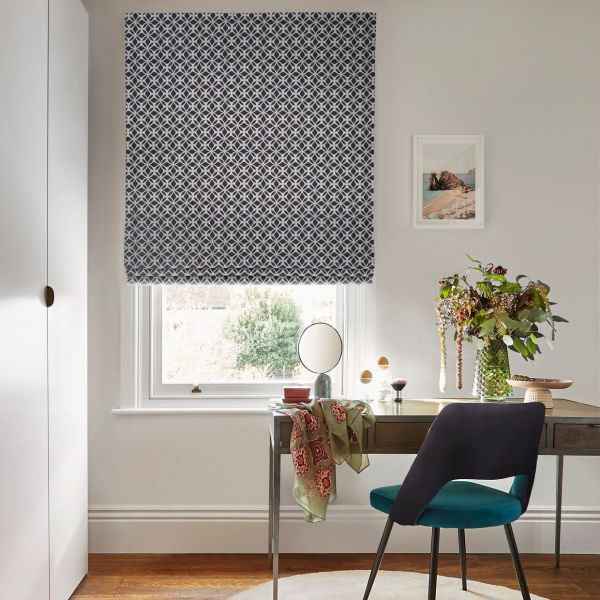 Navy-and-white geometric Roman blind, lowered over most of the window, filtering daylight; a wooden desk beneath in a bright workspace with a teal chair, vase of flowers, and framed print.