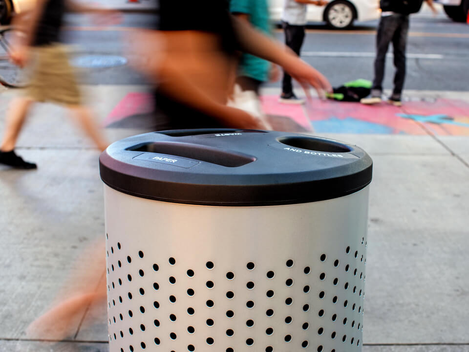 metal powder coated trash and recycling container with a perforated body and black plastic lid placed on a busy city sidewalk
