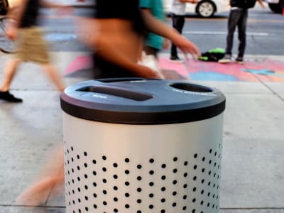 metal powder coated trash and recycling container with a perforated body and black plastic lid placed on a busy city sidewalk