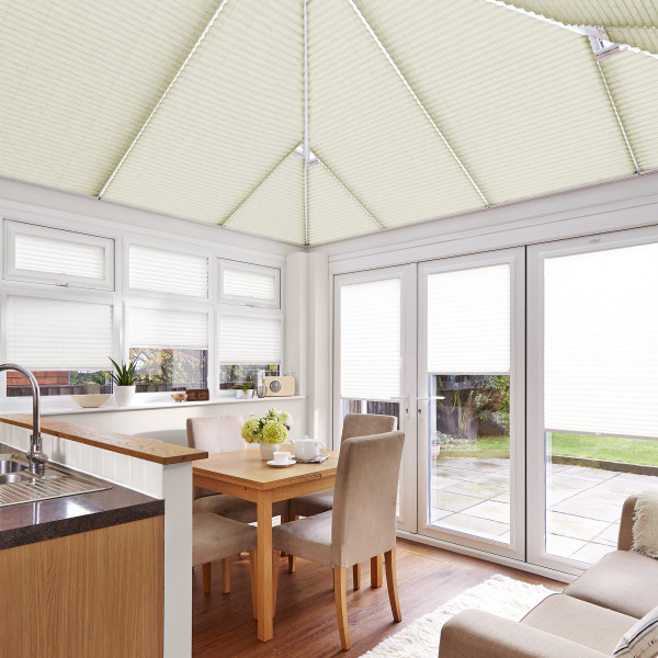 Cream pleated blinds on pitched conservatory roof and over windows and French doors, closed and diffusing bright daylight; small dining area with wooden table, beige chairs, sink and garden view.