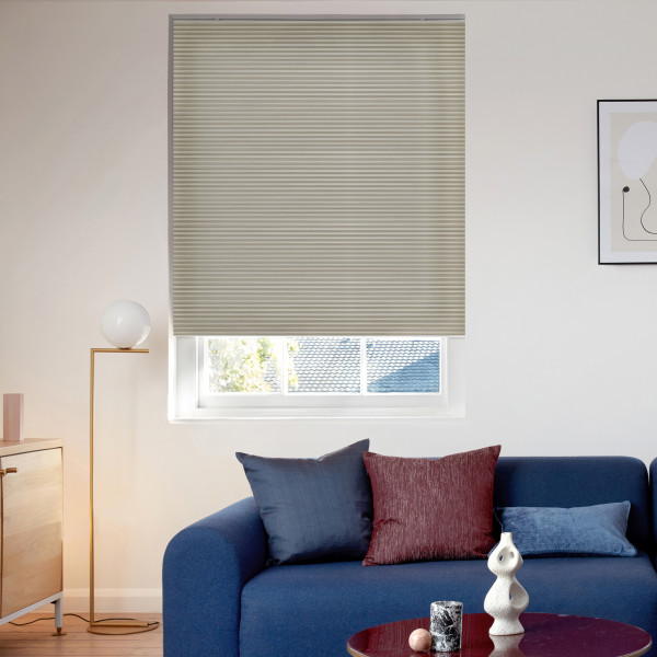 A beige cellular blind pulled down over a recessed rectangular window, closed and filtering daylight into a modern living room with a blue sofa, cushions, floor lamp, and framed art.