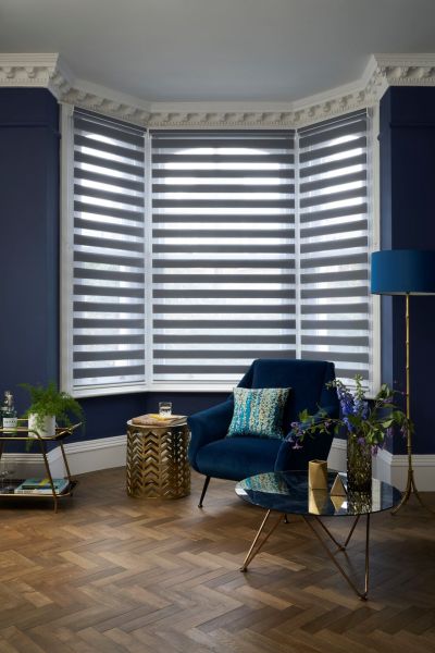 Zebra roller blinds on a three-panel bay window, alternating sheer and opaque stripes, partially closed and diffusing daylight; navy living-room corner with velvet armchair, gold accents, and parquet floor.