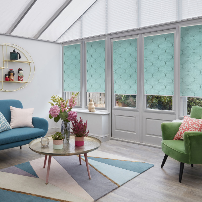 Teal patterned roller blinds cover tall conservatory doors and windows, partially lowered to filter light; white pleated roof blinds above. Sunlit living room with colorful sofas, round coffee table, and flowers.