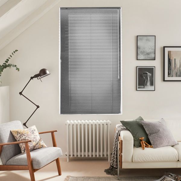 Horizontal aluminum blinds covering a tall rectangular window, fully closed and filtering light. Cozy neutral living room with radiator beneath window, armchair, sofa with cushions, angled floor lamp, wall art.