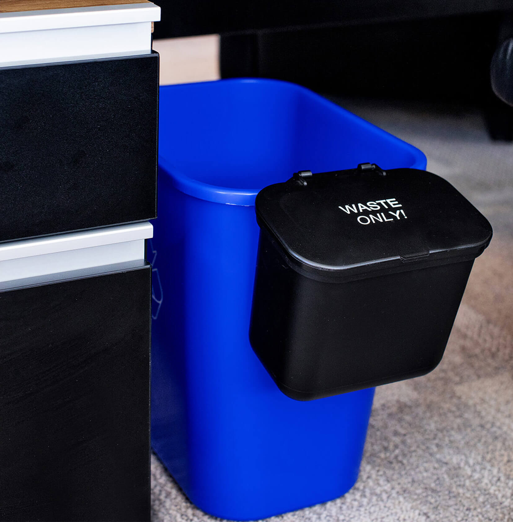 small blue plastic recycling bin with a small black plastic hanging waste container on the side sitting under a desk in an office