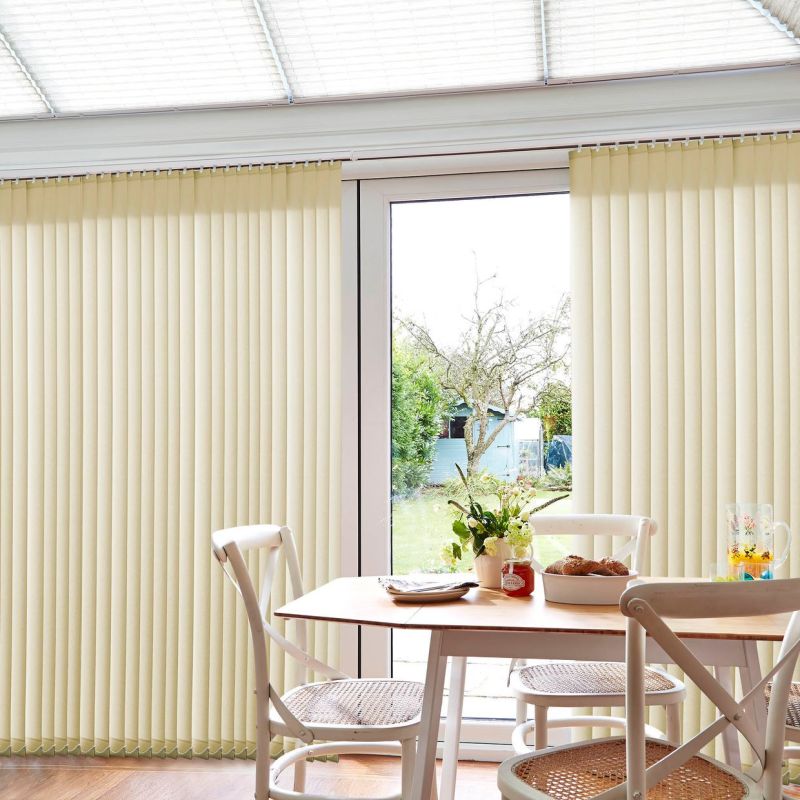 Beige vertical blinds, mostly closed with a central gap, filtering daylight and partly opened to reveal a backyard. Small bright dining area with wooden table, cane‑seat chairs, vase and pastries.