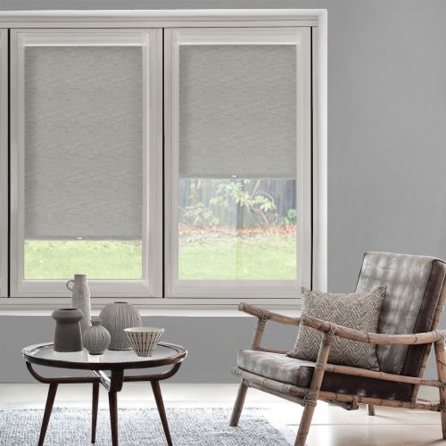 Light-gray fabric roller blinds, partially lowered on twin windows, filtering daylight and leaving lower panes visible; neutral living room with rattan-framed leather chair, patterned cushion, coffee table with vases.