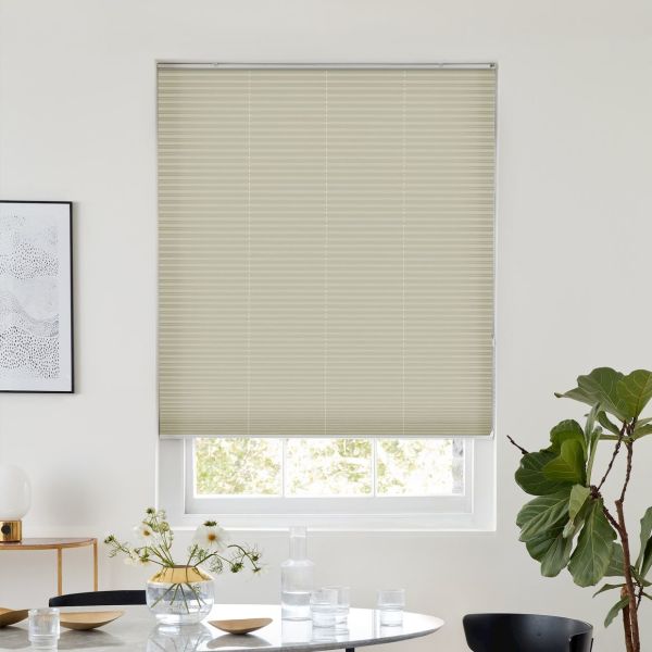 Light-beige cellular blind, fully lowered with horizontal pleats, filtering daylight; modern dining room with round table, vase and glassware, potted fiddle‑leaf fig visible by the window.