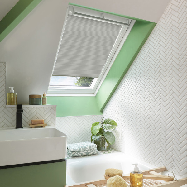 White pleated skylight blind, lowered halfway across the roof window; partly closed, filtering soft daylight; compact attic bathroom with green trim, white herringbone tiles, sink and bathtub.