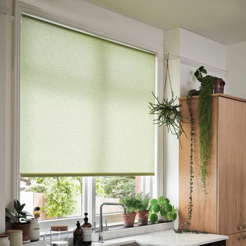 A pale green roller blind lowered over a kitchen window, filtering soft daylight; sink and potted herbs sit on the sill, while a wooden cabinet with trailing plants frames the bright kitchen.