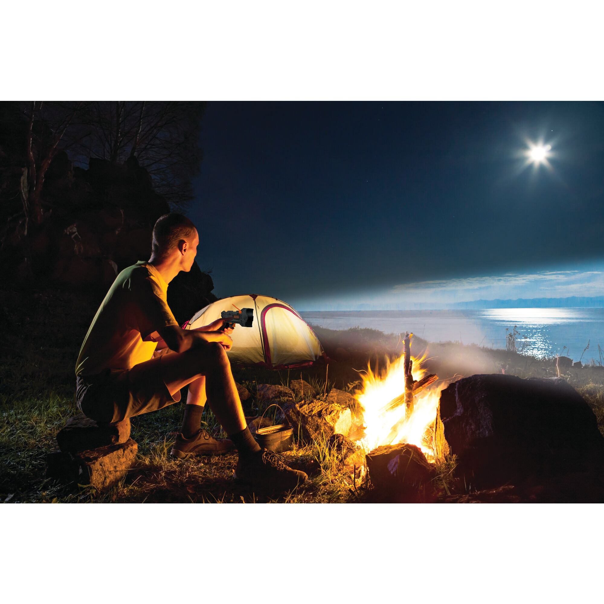 Person camping and sitting near a camp fire using the BLACK+DECKER spotlight