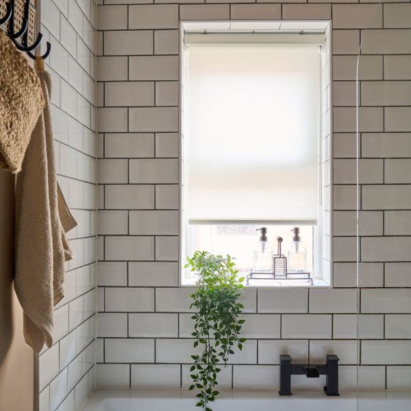 Cream roller blind, fully lowered across window, diffusing bright daylight; trailing potted plant on sill and dual soap dispensers below; white subway-tiled bathroom with black tub faucet and hanging towels.