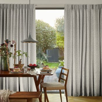 Pleated, floor‑length grey patterned curtains, drawn aside to reveal a sunlit backyard through a large window; wooden dining table set for breakfast, pendant lamp and chairs in the foreground.