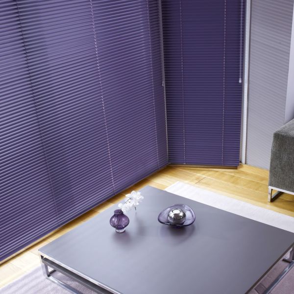 Purple horizontal blinds covering corner floor-to-ceiling windows, tightly closed with visible pull cords; blocking light. Modern living room with low grey coffee table, purple glass ornaments, hardwood floor, grey sofa.