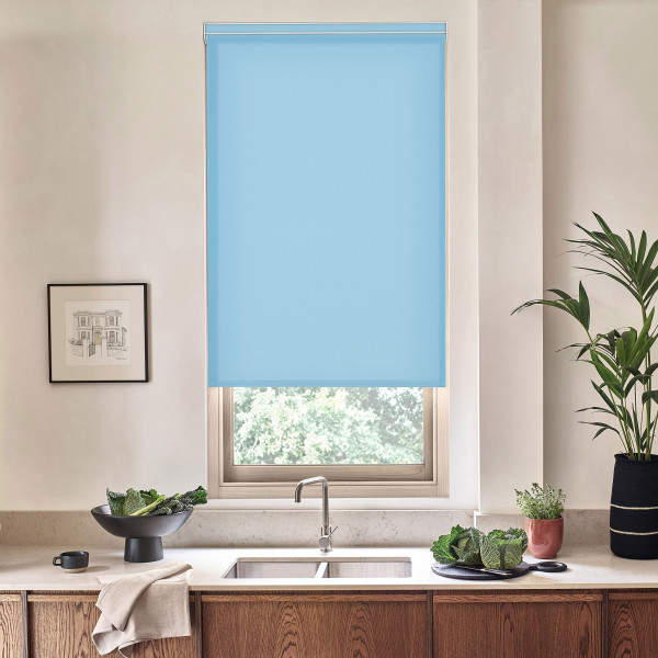 Blue roller blind covers upper portion of a kitchen window; silver faucet and stainless sink below; countertop holds a bowl of leafy greens, potted plants, framed sketch on pale wall.