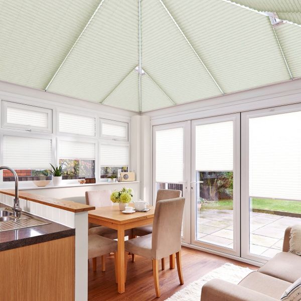 Pale honeycomb/pleated blinds covering the sloped conservatory roof and windows, closed and softly diffusing daylight; small dining area with oak table and beige chairs beside a kitchen counter, patio view.