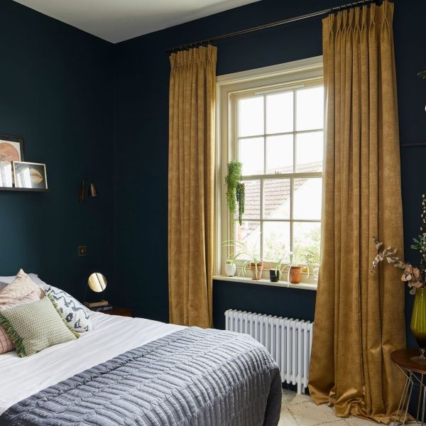 Floor-length mustard velvet pleated curtains, hung on a rod and pulled to the sides, slightly pooling on the floor; framing a sunlit bedroom window with plants, radiator, dark teal walls.