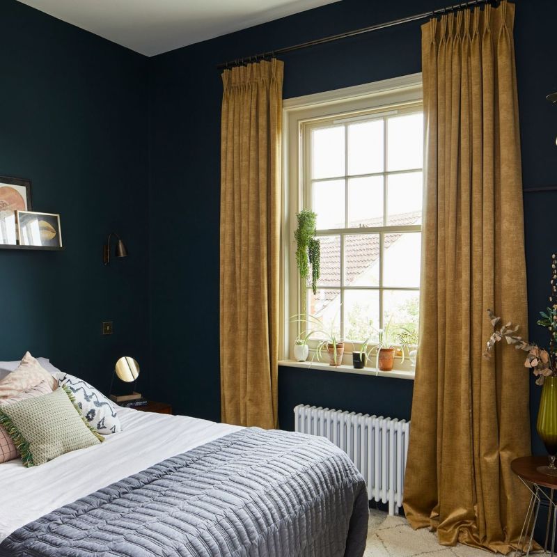 Floor-length mustard velvet pleated curtains, hung on a rod and pulled to the sides, slightly pooling on the floor; framing a sunlit bedroom window with plants, radiator, dark teal walls.
