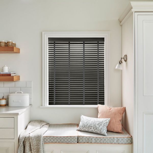 Black horizontal slatted blinds, fully lowered and closed with visible ladder tapes, blocking light; set in a white‑trimmed window above a cushioned window seat in a softly lit neutral kitchen.