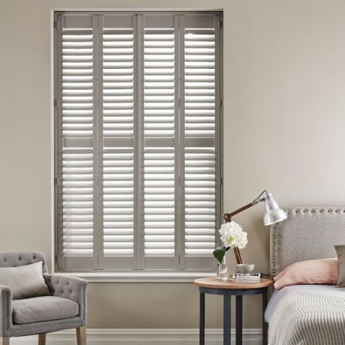 White four-panel louvered plantation shutters covering a tall rectangular window, slats tilted to filter daylight; bedroom setting with upholstered bed, side table with lamp, flowers and a tufted armchair.