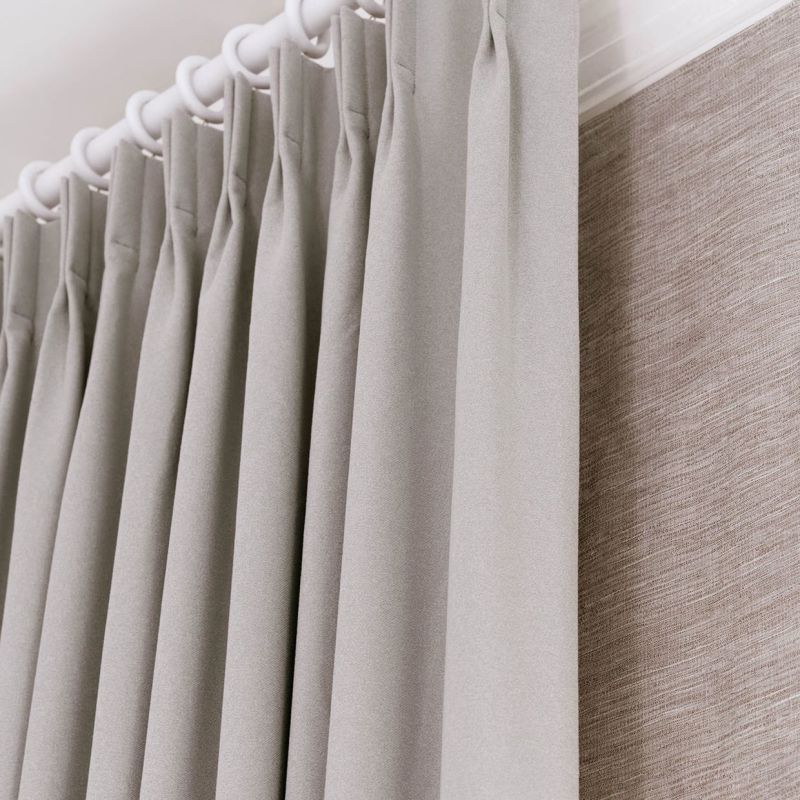Pleated grey fabric curtains hung from a white ring-top rod, tightly closed and evenly pleated; they obscure a textured beige wall covering beside them, bathed in soft, diffused indoor light.