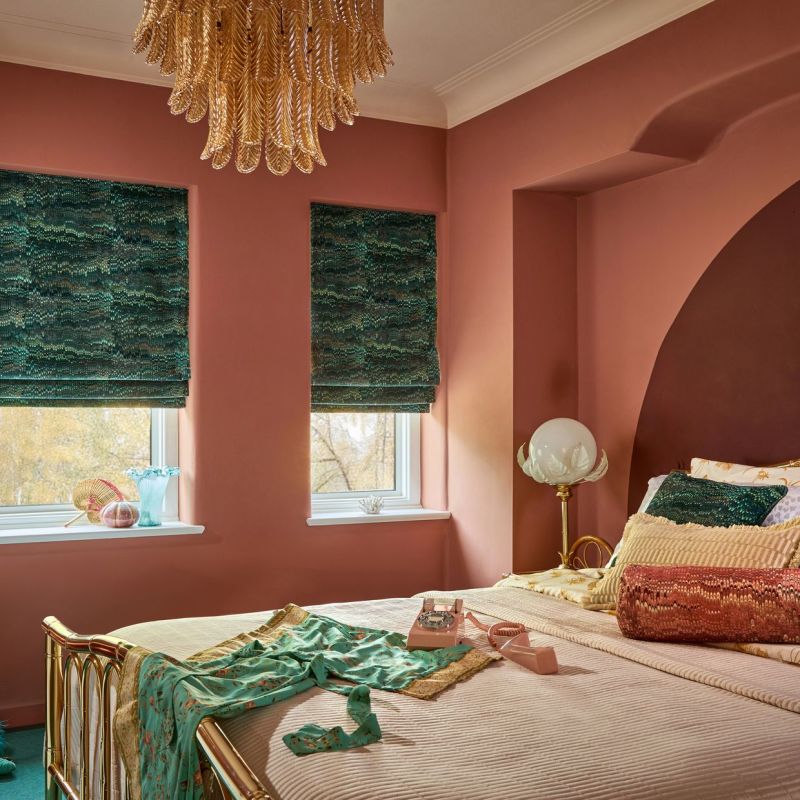 Two green patterned Roman shades, lowered over adjacent windows, filtering soft daylight; set against warm terracotta bedroom walls with brass bed, vintage lamp, layered cushions and leafy outside view.