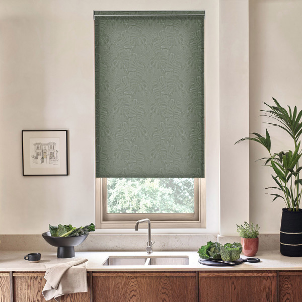 A green embossed roller blind hangs lowered over a window above a stainless sink; a faucet, wooden cabinets, bowls of leafy greens and potted plants sit on the countertop.