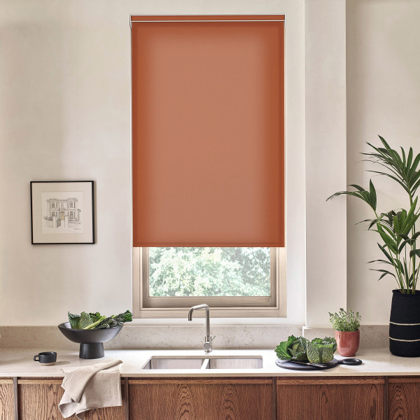 Terracotta roller blind covers a kitchen window, partially blocking daylight; stainless-steel faucet and sink sit below on a marble countertop with wooden cabinets, bowls of leafy greens, and potted plants.