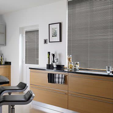 Horizontal aluminum Venetian blinds covering a large window at right and small door window at left, closed and filtering daylight; modern kitchen with light wood cabinets, black countertop, bar stools.