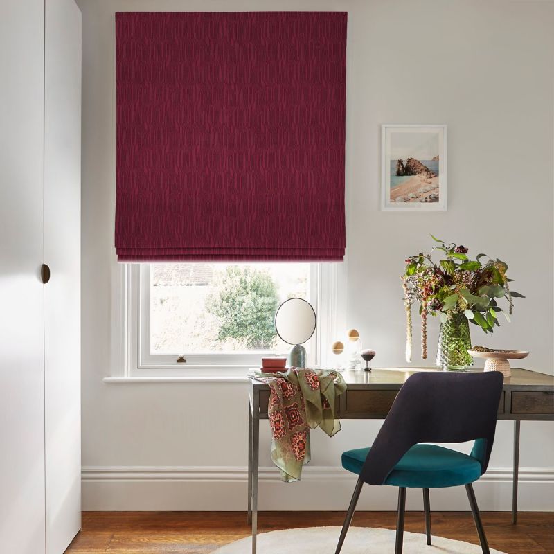 Burgundy patterned Roman blind lowered over a sash window, blocking most light; in a bright home office with a desk, teal-upholstered chair, round mirror, and vase of flowers.