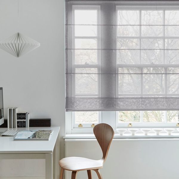 Sheer grey Roman blind, lowered with horizontal folds, filtering pale daylight over a multi‑pane window in minimalist home office with white desk, wooden chair, pendant lamp and bare trees outside.
