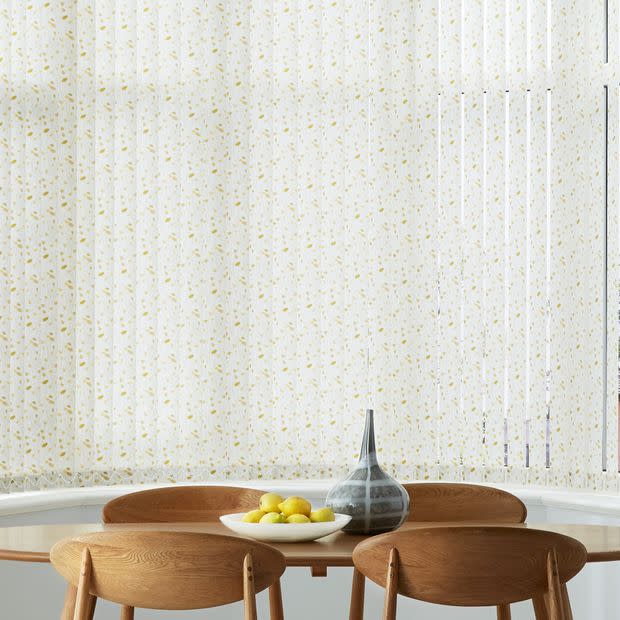 Vertical fabric blinds with small yellow floral print cover a curved bay window, drawn closed and filtering daylight; dining area with round wooden table, bowl of lemons and grey vase.