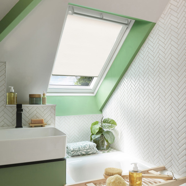 A white pleated skylight blind, partially lowered and filtering daylight over a roof window; set in green-trimmed sloped ceiling above a tiled bathroom with sink, tub, plant, and toiletries.