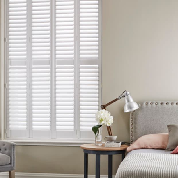White plantation shutters covering a tall window, closed with slatted panels filtering soft daylight; beside a bedside table with lamp, vase, and upholstered headboard in a neutral bedroom.