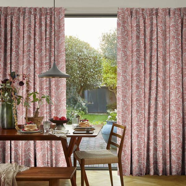 Floor-length pink floral curtains, pulled apart to frame a sliding glass door, admitting daylight into a dining area with a wooden table set for breakfast and a garden beyond.