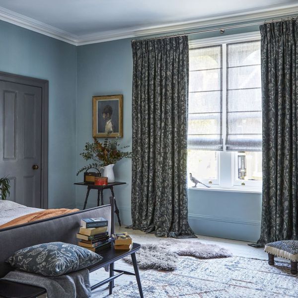 Patterned heavy curtains and sheer Roman blinds â€” curtains parted and pooling at the floor, blinds partially lowered filtering soft daylight â€” in a softly lit blue bedroom with rug, bench, and stacked books.