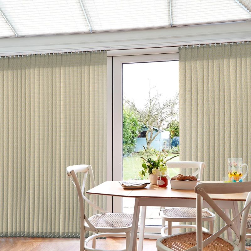 Beige patterned vertical blinds, mostly closed with a central opening, filtering daylight through patio doors; small dining table and chairs in foreground, vase and pastries on table, sunlit backyard beyond.