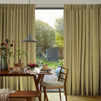 Beige floor-length pleated curtains, slightly parted, framing and filtering sunlight through a sliding glass door; dining table set with pancakes, pendant lamp and wooden chairs, garden view beyond.