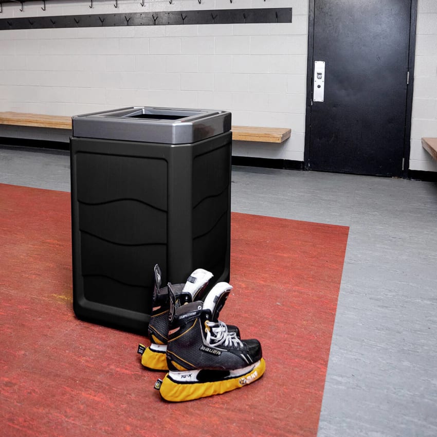 large capacity waste bin with black plastic body and steel look plastic lid in a locker room with a pair of hockey skates sitting on the floor