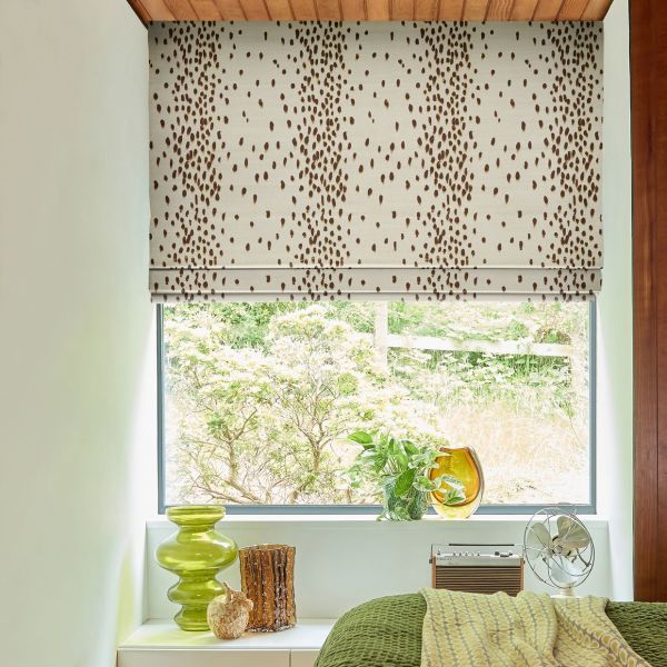 A cream Roman blind with scattered brown spots, lowered halfway and filtering daylight; over a bedroom window with plants on sill, green glass vase, vintage fan and green bedcover.