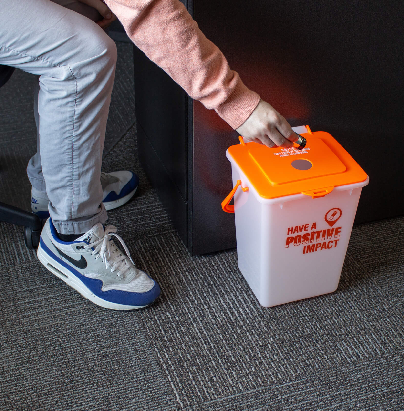 small plastic container with bright orange lid for the collection of used batteries for recycling on the floor with a man inserting used batteries