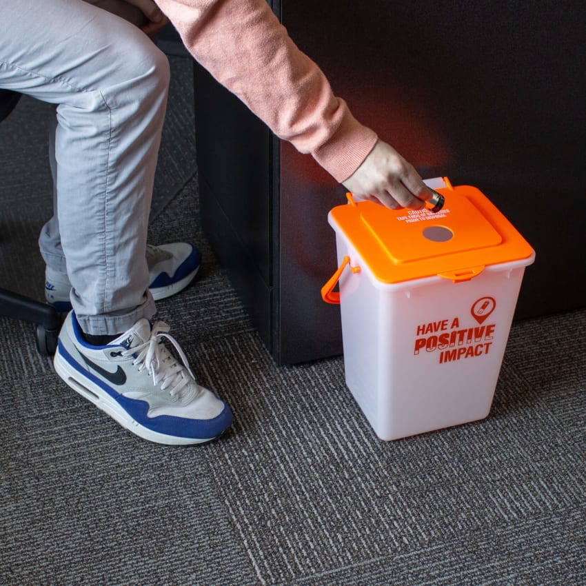 small plastic container with bright orange lid for the collection of used batteries for recycling on the floor with a man inserting used batteries