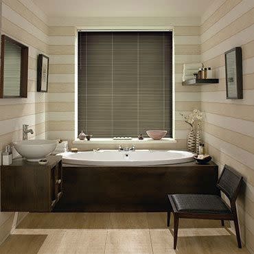 Horizontal Venetian blinds, dark gray, lowered over central window, closed and filtering light; bathroom with white bathtub in a dark wood surround, striped pale walls, vessel sink and chair.