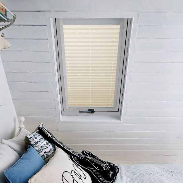 Beige pleated skylight blind, fully closed and filtering daylight, set inside a white-framed roof window; attic bedroom with white painted wooden walls and a bed piled with patterned cushions.