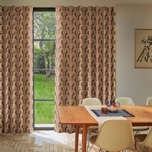 Patterned floor-to-ceiling pleated curtains, pink geometric print, drawn across a tall multi-pane window, filtering daylight into a dining room with a wooden table, chairs, and a grassy garden view.