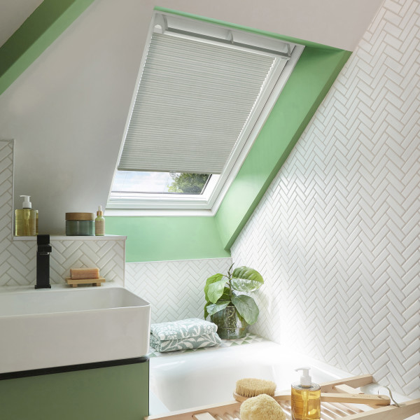 White pleated skylight blind, partially lowered over an angled roof window, filtering soft daylight; set in a bright attic bathroom with mint-green trim, white herringbone tiles, sink, tub, plant.