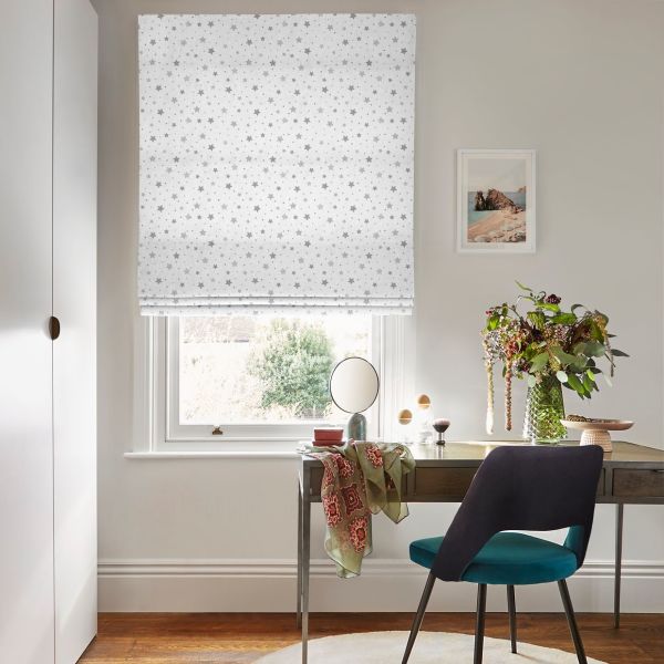 White Roman blind with small grey stars, lowered and filtering light; over a desk with teal chair, vase of flowers in a bright home office with framed seaside print.
