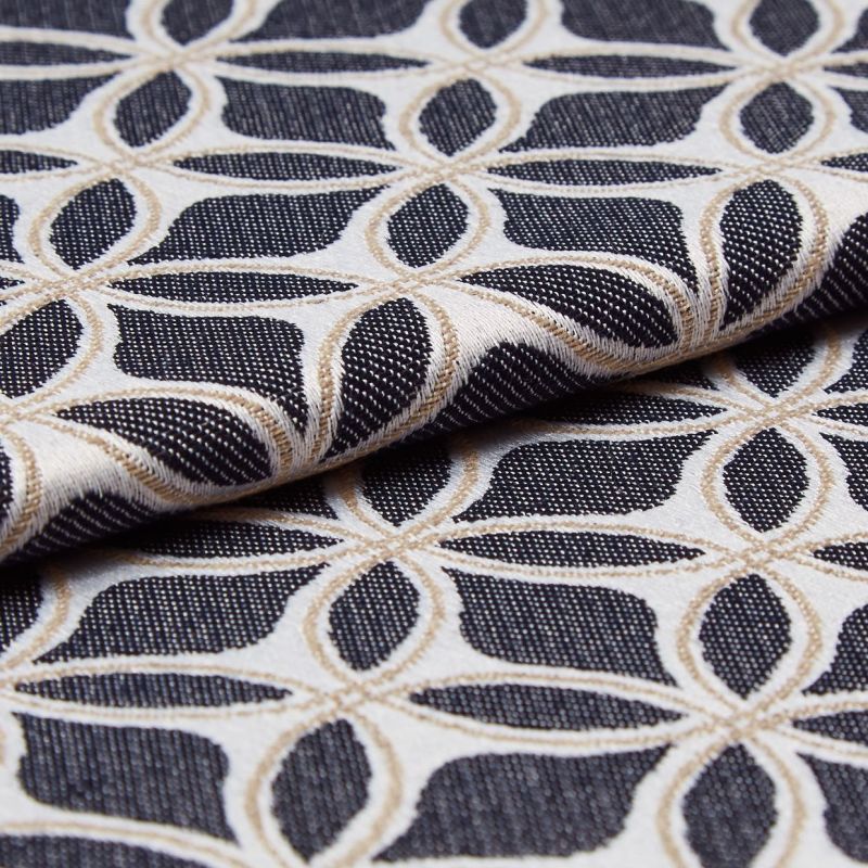 Jacquard upholstery fabric with cream and tan interlocking petal lattice on a black textured ground, folded across itself, showing raised embroidered weave; close-up, softly lit studio setting.