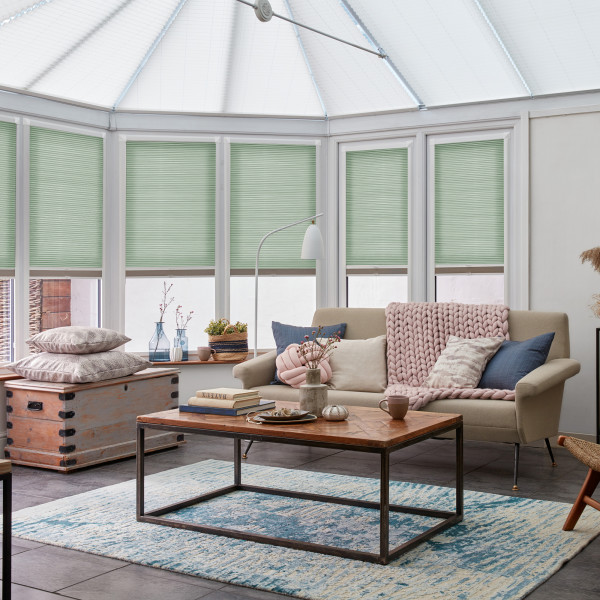 Light-green pleated cellular blinds lowered halfway across tall bay windows, filtering soft daylight; conservatory-style living room with beige sofa, wooden coffee table, blue rug, floor lamp and decorative cushions.